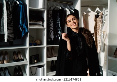 Stylish Female Smiling And Looking At Camera While Choosing Outfit In Modern Walk In Closet With Various Clothes And Heels At Home