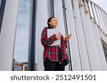 A stylish female student in a red shirt with a laptop who looks at the schedule of lessons on the phone standing against the background of the university.