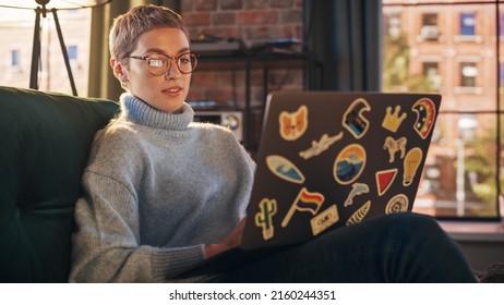 Stylish Female Using Laptop Computer With Diverse LGBT And Lifestyle Stickers On The Back. Young Creative Woman Sitting On A Couch, Typing, Browsing Internet And Checking Social Media At Home.