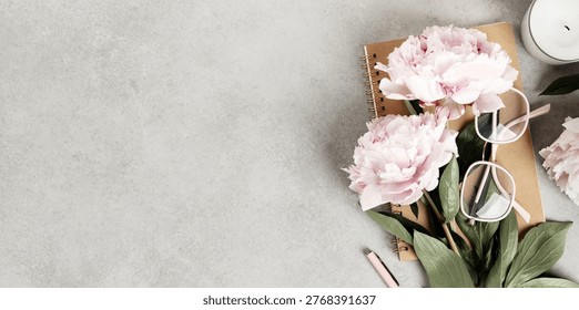 Stylish flat lay composition featuring pink peonies, green leaves, a spiral notebook, eyeglasses, and a pen on a textured gray background. Feminine and creative workspace vibe.