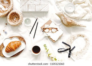 Stylish flat lay of workspace in bed with cards,wooden box,croissant, glasses,cotton baskets, woman accessories on white bedsheets background.Flat lay, top view blogger influencer home office concept.