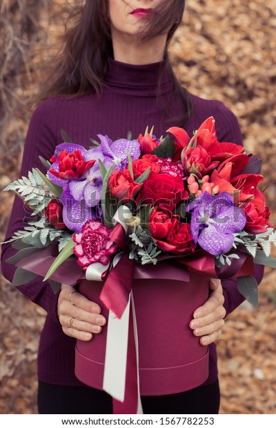 Stylish flower\
arrangement in a burgundy hatbox. Purple Orchodia vanga, red\
Amaralis and tulips with bunch. The girl holds the flower bouquet\
in her hands. Birthday\
present.