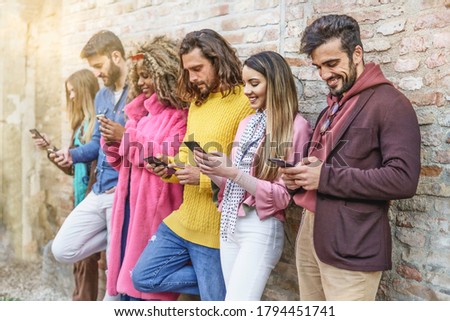 Stylish friends having fun watching smart phones screen against a wall. Happy young people staying online using the internet and social media apps.