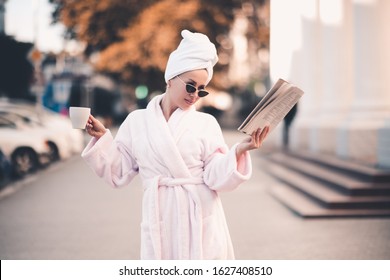 Stylish girl 24-26 year old wearing white bathrobe and towel, reading fresh magazine and drinking tasty coffee standing outdoors on street closeup. Good morning. Spa resort. 