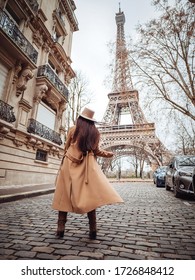 A stylish girl in a beige coat, hat and boots stands on a small Parisian street with a view of the Eiffel Tower. Paris style.