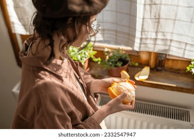 Stylish girl in beret peels orange near windowsill with houseplants at home. Sunny day in harmony with yourself. Artist woman holding fresh fruit in hand enjoy fragrance pondering new idea for painted - Powered by Shutterstock