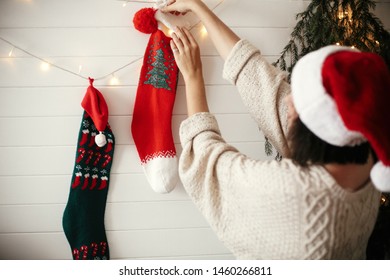 Stylish girl in cozy sweater and santa hat decorating room for christmas holidays with stockings, garland light and christmas tree on white wall . Merry Christmas. Happy Holidays - Powered by Shutterstock