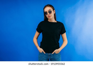 Stylish girl in glasses wearing black t-shirt posing in studio on blue background