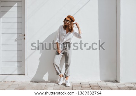 Stylish girl in gray pants and white cotton blouse posing near white wall. Photo of woman in cap and glasses
