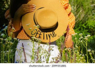 Stylish girl holding a straw hat with a words Offline. A concept of summer vacation away from a big city, human unity with nature. Woman standing in a blooming meadow among wildflowers on a sunny day