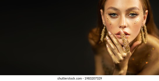 Stylish girl looks at camera. Gold ring on finger, earrings rings. Beautiful face, blue eyes, blonde hair, golden glamorous makeup, gold paint on body, skin, hands. Portrait of fashion model woman