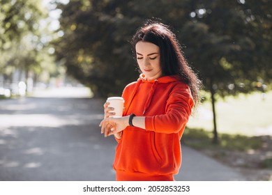 Stylish Girl In Red Sweater Looking At The Watch And Drinking Coffee. Happy Woman Is Waiting Friends And Relaxing In The Park, Outdoors, Summer Time