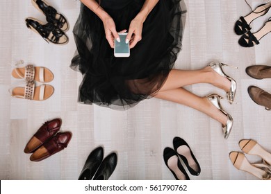 Stylish girl sitting on the floor in a dressing room with smartphone in hands, writes  the message, surrounded by a variety of shoes. She is dressed in a black skirt, on her feet silver luxury shoes  - Powered by Shutterstock