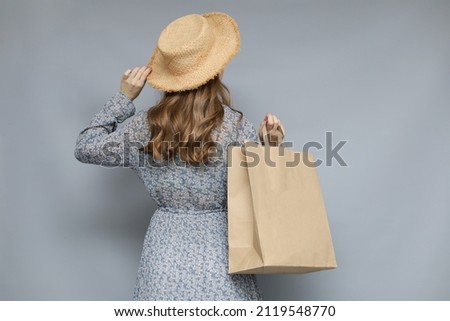 Stylish girl in straw hat holds brown blank paper bag. Studio shot with space for logo or design. Picture taken from the back.