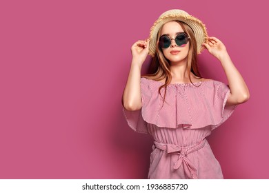 Stylish girl in striped dress standing in straw hat and big round glasses on pink background