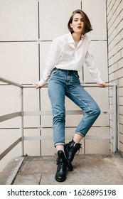 Stylish girl in vintage jeans and white cotton blouse posing near wall. Photo of woman outfit. Short haircut