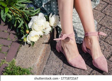 A stylish girl's legs in fashionable pink flat shoes with ankle ties and sharp toe sitting on the brick pavement with white peony flowers and a light green skirt on a sunny day in Amsterdam