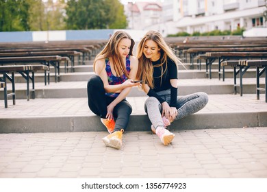 Stylish girls in a sportwear. Two woman sitting in a sunny park. Girls use the phone 