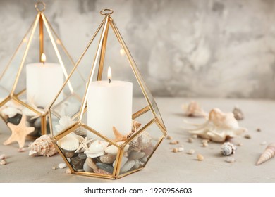 Stylish glass holders with burning candles, seashells and pebbles on light stone table