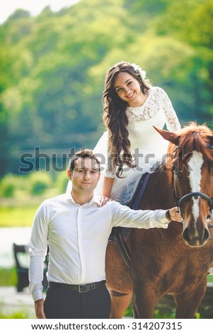stylish gorgeous happy brunette bride riding a horse and elegant groom on the background of a park