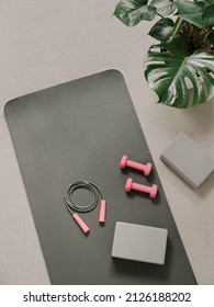 Stylish gray and pink home fitness flat lay. Top view of gray sport mat, yoga block, skipping rope and pink dumbbells on neutral carpet background, monstera plant. Set for pilates, fitness, yoga