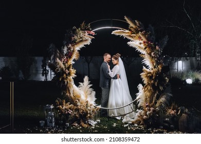 A stylish groom and a beautiful bride in a long dress are standing at night near a reed arch decorated with lamps and garlands. Wedding photography, portrait.