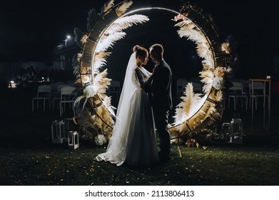 A stylish groom and a beautiful bride in a long dress are standing, hugging at night near a luminous cane arch decorated with lamps and garlands. Wedding photography, portrait.