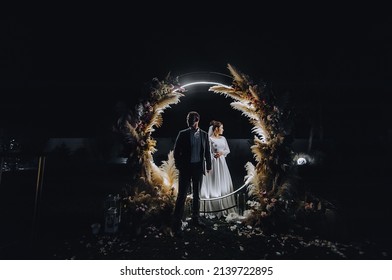 A Stylish Groom And A Beautiful Bride In A Long Dress Are Standing At Night Near A Reed Arch Decorated With Lamps And Garlands. Wedding Photography, Portrait.