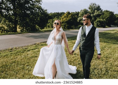 Stylish groom and beautiful young bride in a white dress are walking holding hands in nature outdoors at sunset. Wedding photography, portrait of smiling newlyweds in sunglasses. - Powered by Shutterstock