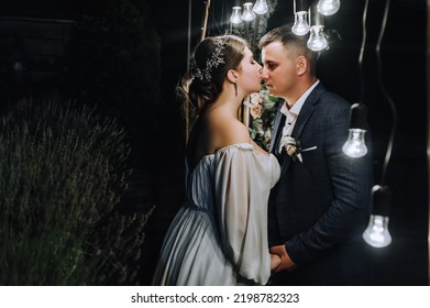 A Stylish Groom In A Blue Suit And A Beautiful Bride In A White Dress Hug, Kiss Near An Arch Decorated With Flowers And Lamps At A Ceremony At Night. Wedding Photography, Portrait Of The Newlyweds.