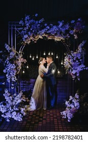 A Stylish Groom In A Blue Suit And A Beautiful Bride In A White Dress Hug Near An Arch Decorated With Flowers And Lamps At A Ceremony At Night. Wedding Photography, Portrait Of The Newlyweds.