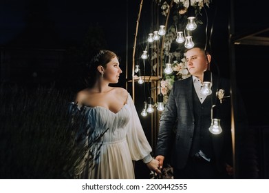 A Stylish Groom In A Blue Suit And A Beautiful Bride In A White Dress Stand Holding Hands Near An Arch Decorated With Flowers And Lamps At A Ceremony At Night. Wedding Photography, Portrait.