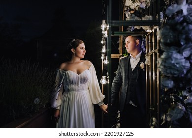 A Stylish Groom In A Blue Suit And A Beautiful Bride In A White Dress Stand Near An Arch Decorated With Flowers And Lamps At A Ceremony At Night. Wedding Photography, Portrait Of The Newlyweds.