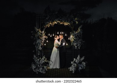 A Stylish Groom In A Blue Suit And A Beautiful Bride In A White Dress Stand Near An Arch Decorated With Flowers And Lamps At A Ceremony At Night. Wedding Photography, Portrait Of The Newlyweds.