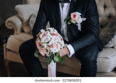 A stylish groom in a blue suit with a boutonniere with a beautiful bouquet in his hands sits on a sofa in a room indoors. Wedding photography, portrait.