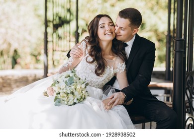 Stylish groom in a gray checkered suit and a cute blonde bride in a white lace dress with a long veil are hugging in the park near the old house. Wedding portrait of smiling newlyweds. - Powered by Shutterstock