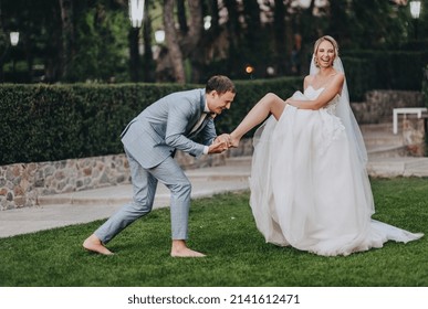 Stylish groom in a gray checkered suit kisses the leg of the blonde bride in a long white dress in a meadow with green grass, against the background of the forest. A funny and funny photo.
