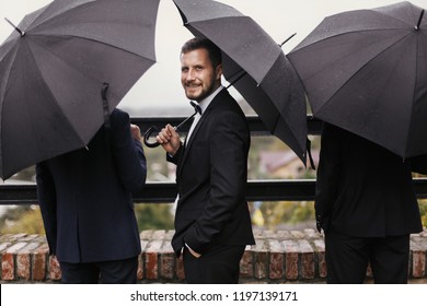 Stylish Groom And Groomsmen Standing Under Black Umbrella And Posing. Confident Man In Suit Holding Umbrella In Rainy Outdoors. Rich Businessman. Finance Wealth And Business