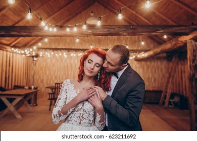 Stylish Groom And Happy Bride Gently Hugging Under Retro Bulbs Lights In Wooden Barn. Rustic Wedding Concept, Space For Text. Newlyweds Couple Embracing, Sensual Romantic Moment