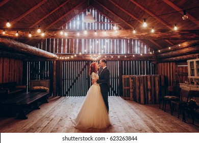 stylish groom and happy bride hugging under retro bulbs lights in wooden barn. rustic wedding concept, space for text. newlyweds couple embracing, sensual romantic moment