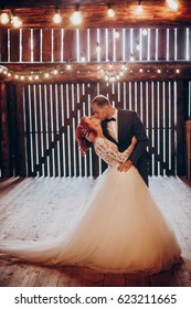 stylish groom and happy bride kissing under retro bulbs lights in wooden barn. rustic wedding concept, space for text. newlyweds couple embracing, sensual romantic moment