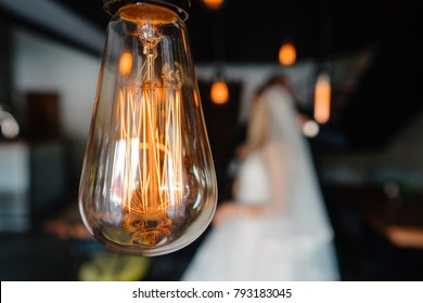 Stylish Groom And Happy Bride Kissing Under Retro Bulbs Lights In Wooden Barn. Rustic Wedding Concept, Space For Text. Newlyweds Couple Embracing, Sensual Romantic Moment