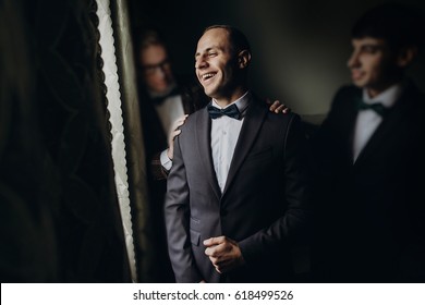 Stylish Groom Laughing And Having Fun With Groomsmen While Getting Ready In The Morning For Wedding Ceremony. Luxury Man In Suit With His Friends Smiling In Room. Space For Text.