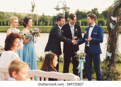 Stylish groom shakes hands with groomsmen standing before wedding altar - Powered by Shutterstock