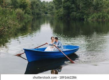Stylish groom in a suit and a beautiful bride in a white lace dress are sitting in a wooden boat and swimming on the lake, enjoying nature.