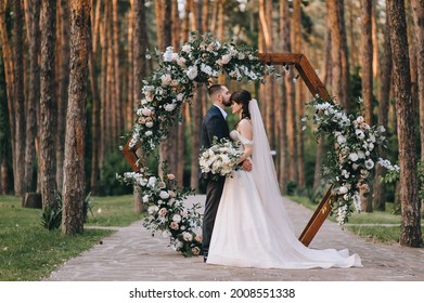 Stylish Groom In A Suit And A Cute Brunette Bride In A White Dress In The Forest Near A Wedding Wooden Arch Decorated With Flowers. Wedding Portrait Of The Newlyweds.