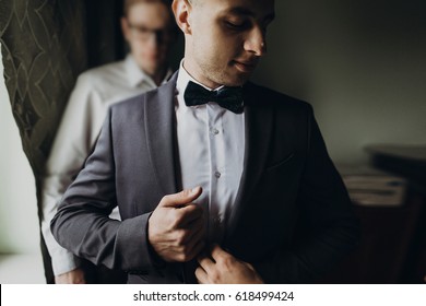 Stylish Groomsmen Helping Happy Groom Getting Ready In The Morning For Wedding Ceremony. Luxury Man In Suit In Room. Space For Text. Wedding Day.