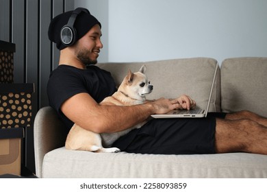 A stylish guy dressed in black clothes, with headphones, at home with his dog, work on a laptop, play board games, spend time together. The guy works on-line, makes purchases on a laptop, IT - Powered by Shutterstock