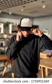 Stylish Guy Looking Down While Posing In Custom Apparel, Black Baseball Cap And Sweatshirt. Young Man Working At Custom T-shirt, Clothing Printing Company. Vertical Shot