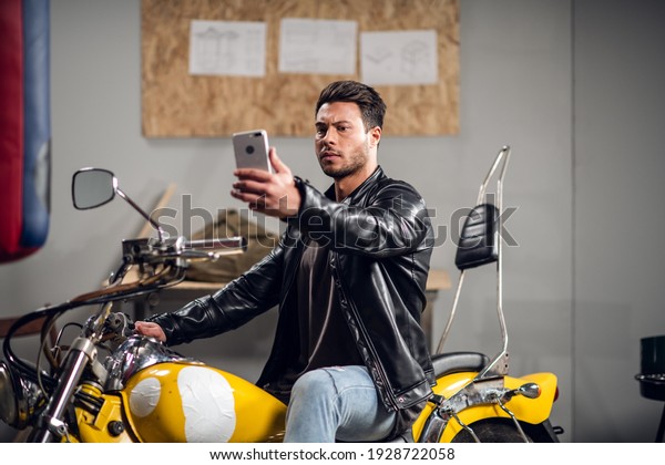 A stylish guy on a yellow cool\
motorcycle takes a selfie in a garage or car repair\
shop.